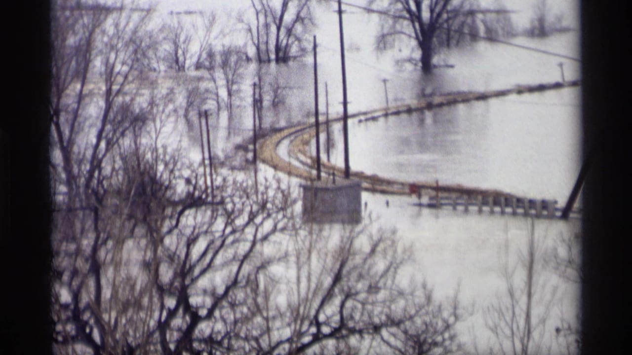 1969 Savage, Minnesota flood view through a dark window frame, showing water around a curving rail embankment and leafless trees.