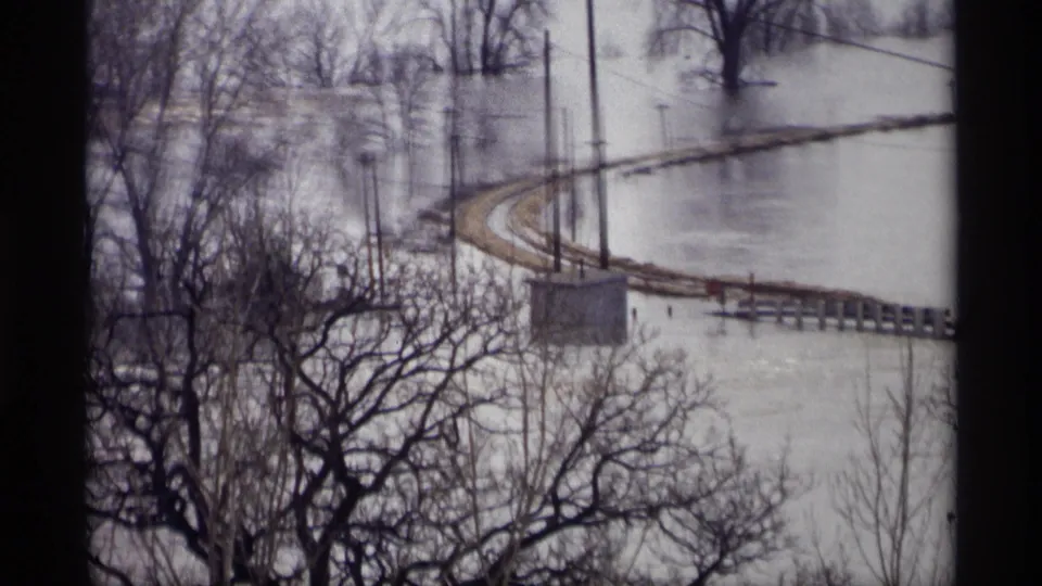 1969 Savage, Minnesota home-movie frame of floodwater, a raised curving rail bed, bare trees, poles, and a dark interior window frame.