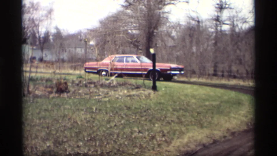 1969 Savage, Minnesota frame of the red sedan moving rightward around a muddy curve near a grassy yard and bare trees.