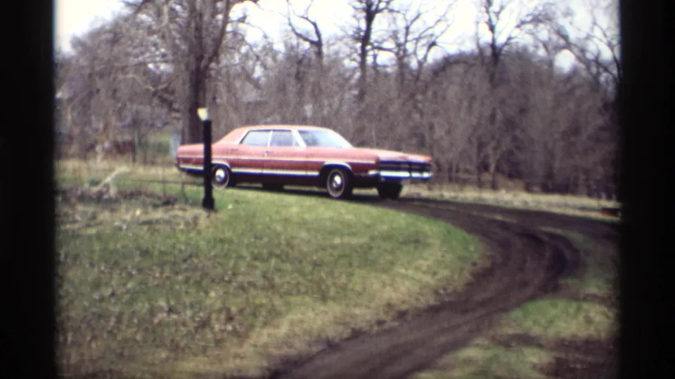 The red four-door sedan appears closer as it follows the muddy curve through the Savage, Minnesota yard setting in 1969 footage.