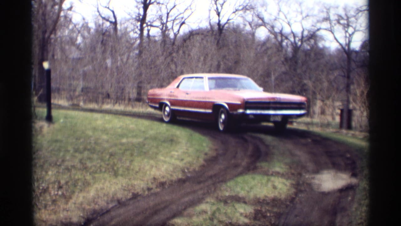 Red classic sedan turning onto a muddy driveway bordered by grass and bare trees.