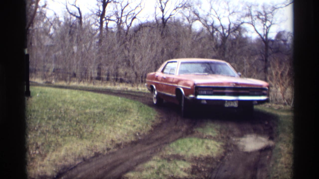 Red sedan closer on the muddy track with grass and bare trees around the drive.