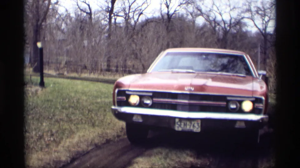 The red sedan fills the frame near the camera, headlights lit and front plate visible on the muddy Savage driveway.