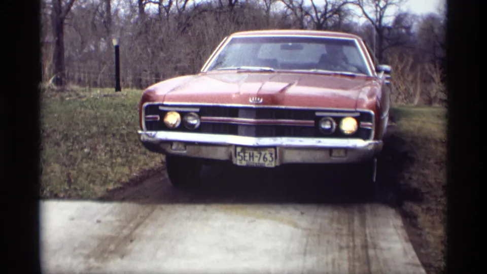 Red Ford car in the supplied 1969 Savage, Minnesota setting, shown front-on with headlights lit beside a grassy rural drive.