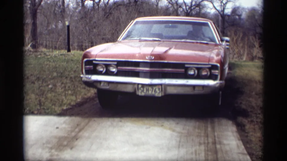 Front view of a red Ford car in 1969 Minnesota home-movie footage, with the grille, plate, driveway, grass, and bare trees visible.