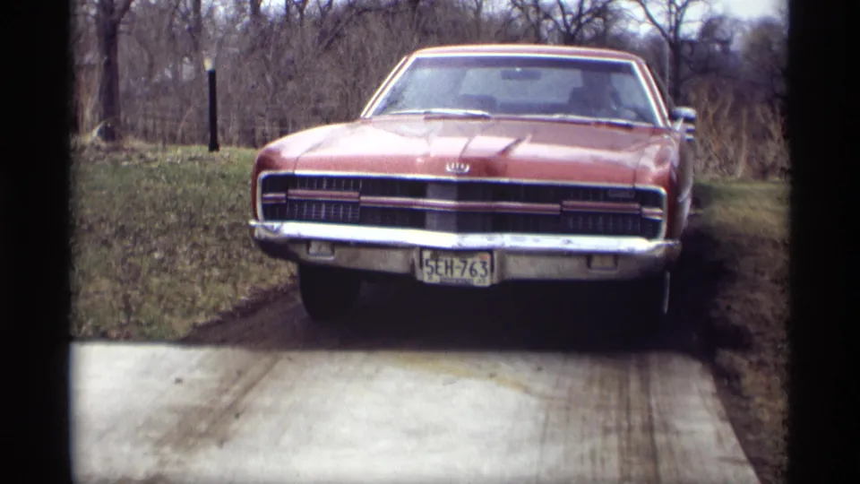 Red Ford car parked on a driveway in the supplied Savage, Minnesota 1969 home-movie scene, with rural yard and bare trees behind it.