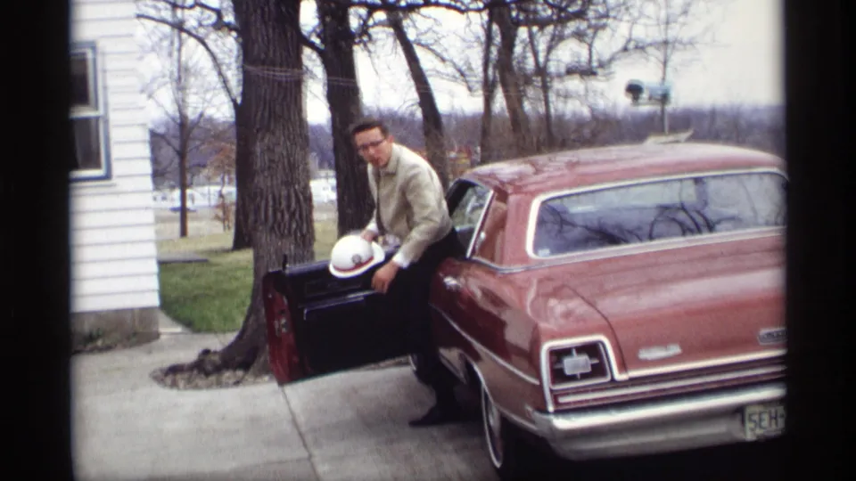 In Savage, Minnesota, 1969, a man in a light jacket and glasses exits a maroon sedan with a white hard hat in hand.