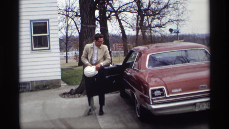 The man stands in the driveway beside the open door of the maroon sedan, holding a white hard hat near a white house.