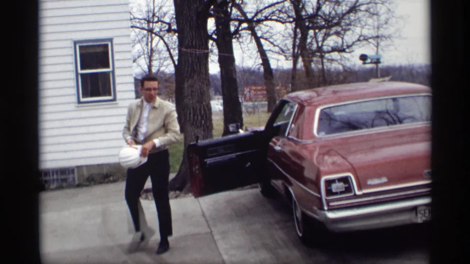 A man in a light suit walks along the driveway with a white hard hat as the maroon sedan remains parked behind him.