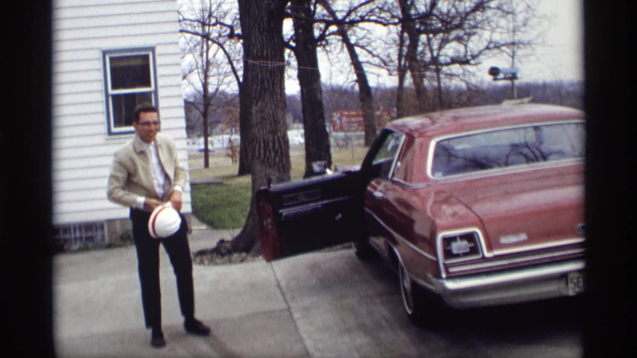 In Savage, Minnesota in 1969, a man stands near an open car door beside a red sedan parked by a white-sided house.