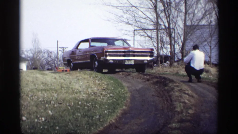 A red sedan sits on a grassy slope in Savage, Minnesota, with a man in a white shirt crouching near the dirt path.