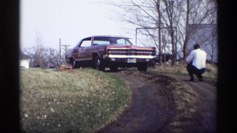 The 1969 home-movie frame shows the red sedan on a sloped yard with bare trees and a man crouched near the driveway edge.