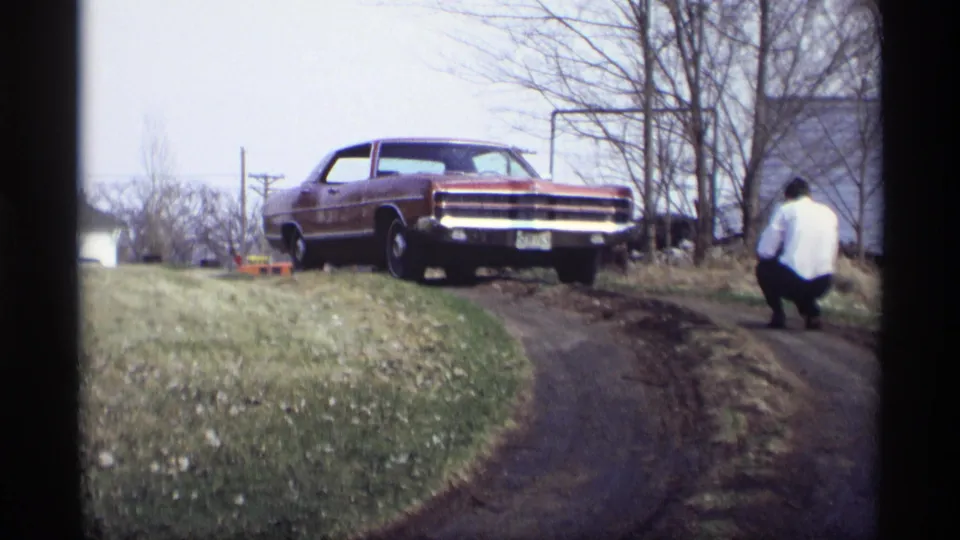 A red 1960s sedan is parked above a curved dirt path in Savage, Minnesota, while a man crouches beside the slope.