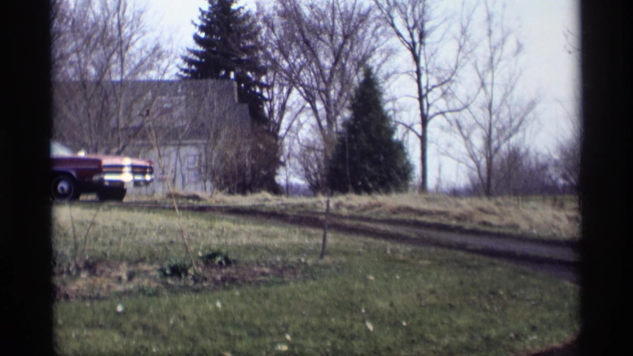 Maroon sedan near a house and bare trees beside a grassy dirt road.