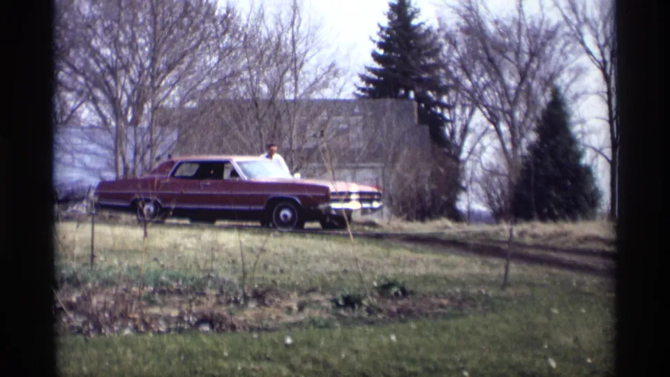 A man in a light shirt stands near a maroon sedan on a grassy hill in Savage, Minnesota, with a house and bare trees behind.