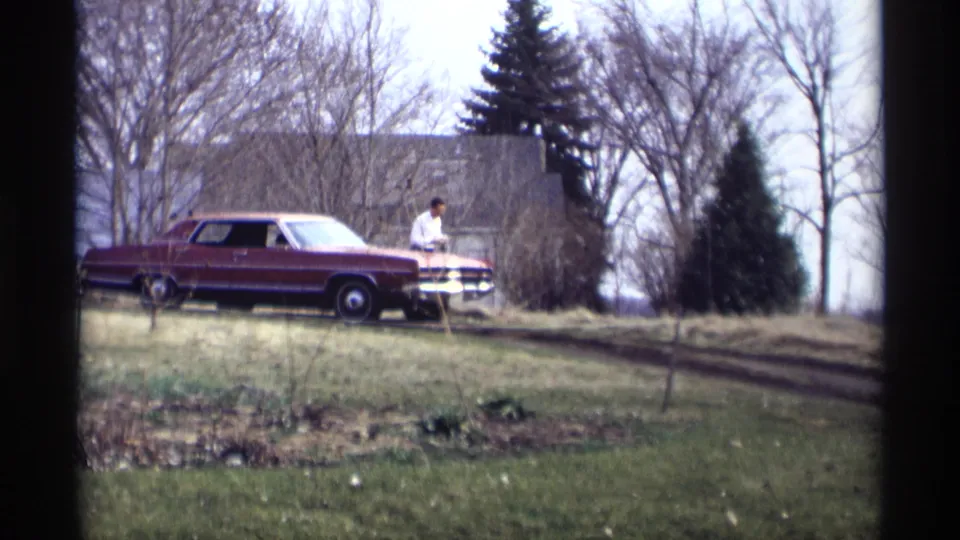 The man remains near the front of the maroon sedan in this 1969 Savage, Minnesota frame, with trees and a house in the background.