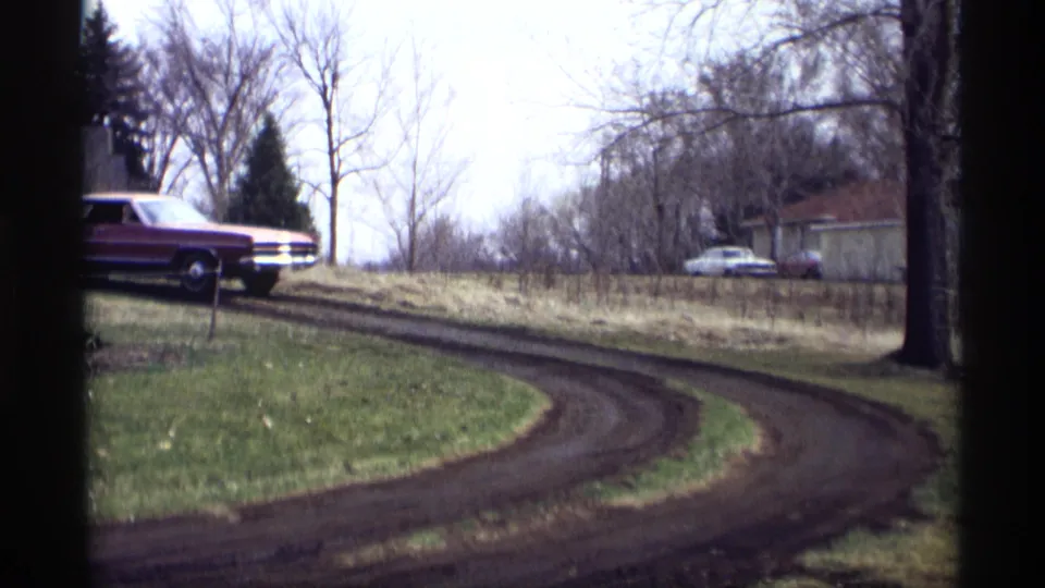 The maroon sedan begins along the curving dirt road in Savage, Minnesota, with a second car and outbuildings visible in the distance.