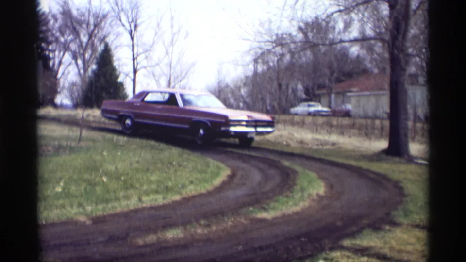 The maroon sedan continues around the dirt driveway bend in Savage, Minnesota, with bare trees and a small building in the background.