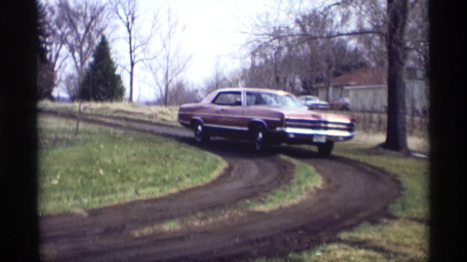 A side view shows the maroon sedan negotiating a sharp muddy driveway turn in this 1969 Savage, Minnesota home-movie frame.