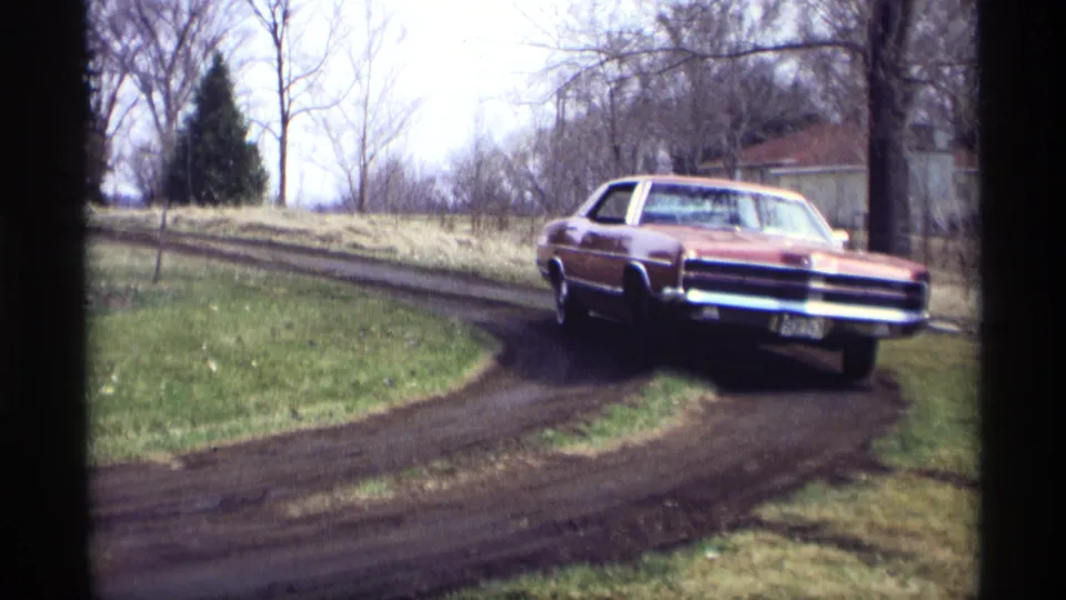 The sedan approaches the camera on the curved dirt driveway, with grass, bare trees, and a small background structure visible.