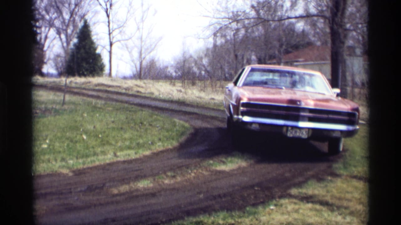In Savage, Minnesota 1969 footage, a red 1960s sedan approaches along a muddy curved road near grass, bare trees, and a distant building.
