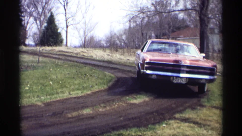 In Savage, Minnesota 1969 footage, a red 1960s sedan approaches along a muddy curved road near grass, bare trees, and a distant building.