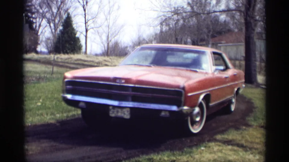 The red 1960s sedan nears the camera on the muddy Savage, Minnesota road, with chrome trim, grille, and a whitewall front tire visible.