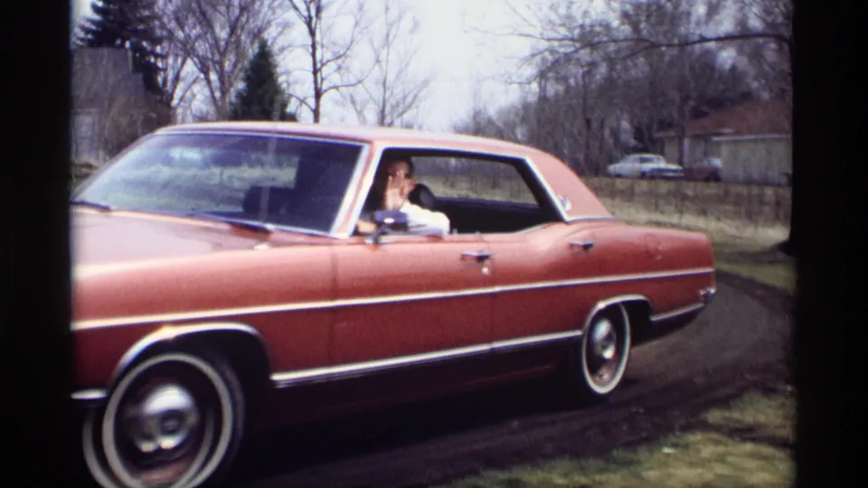 A red 1960s sedan passes close by in Savage, Minnesota, with the driver visible at the open side window and whitewall tires below.