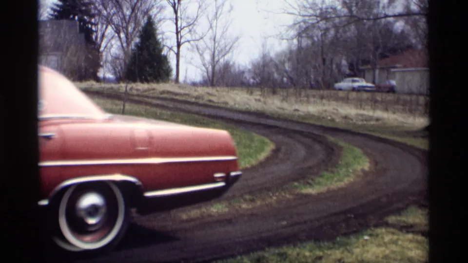 The red sedan continues out of frame along the muddy curved road in Savage, Minnesota, leaving the rural roadside and distant house visible.