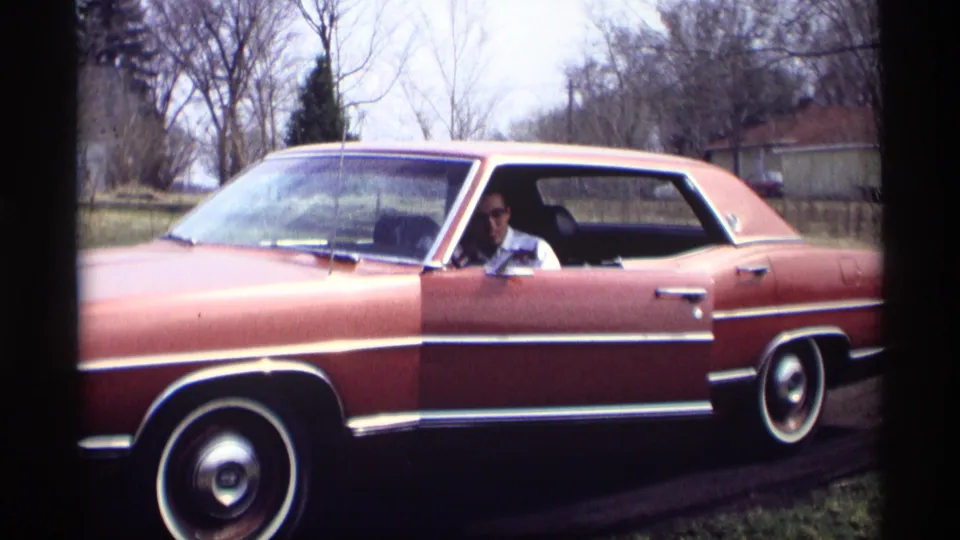 In Savage, Minnesota home-movie footage from 1969, an adult man sits inside a reddish-brown sedan near bare trees and a roadside.