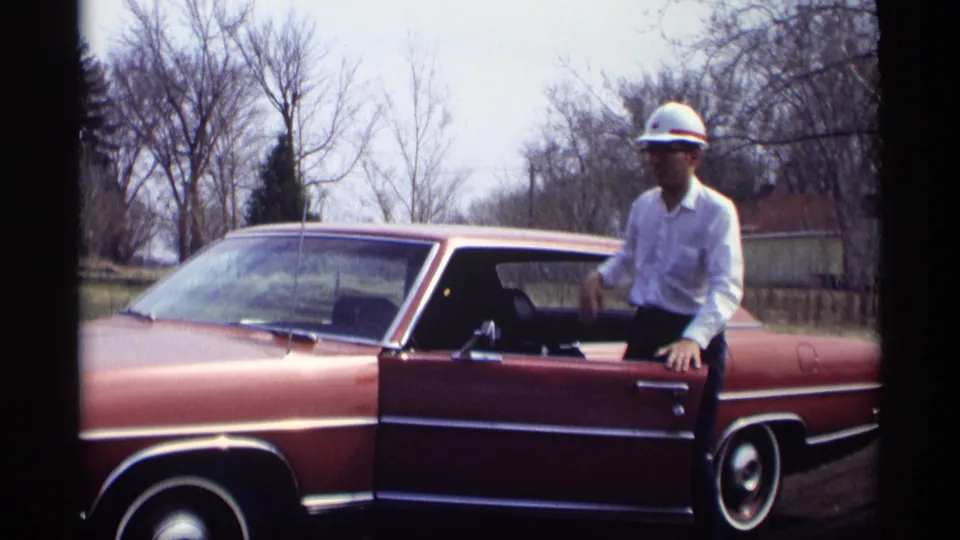 The man now wears the white hard hat and stands by the open sedan door in the 1969 Savage, Minnesota roadside scene.