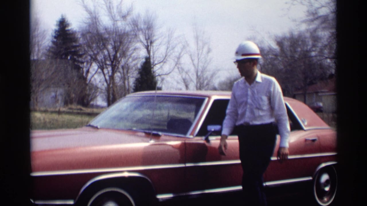Adult man in a white hard hat and light shirt stands near a red sedan in 1969 Minnesota home-movie footage.