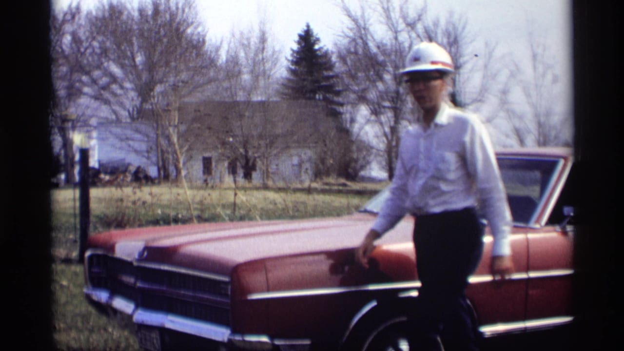Man in a hard hat walks near the front of a red sedan outdoors.