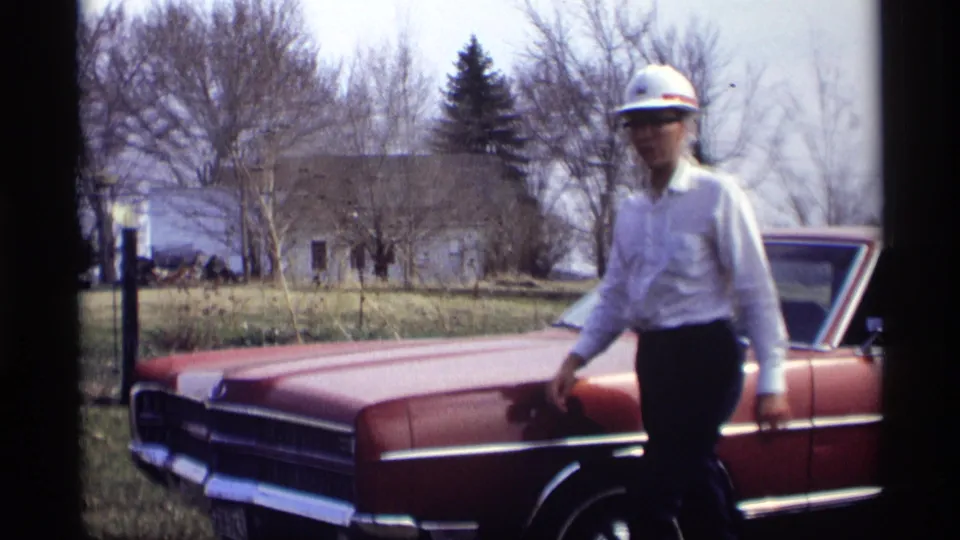 The hard-hat man moves past the front of a red sedan, with a yard and bare trees behind him.
