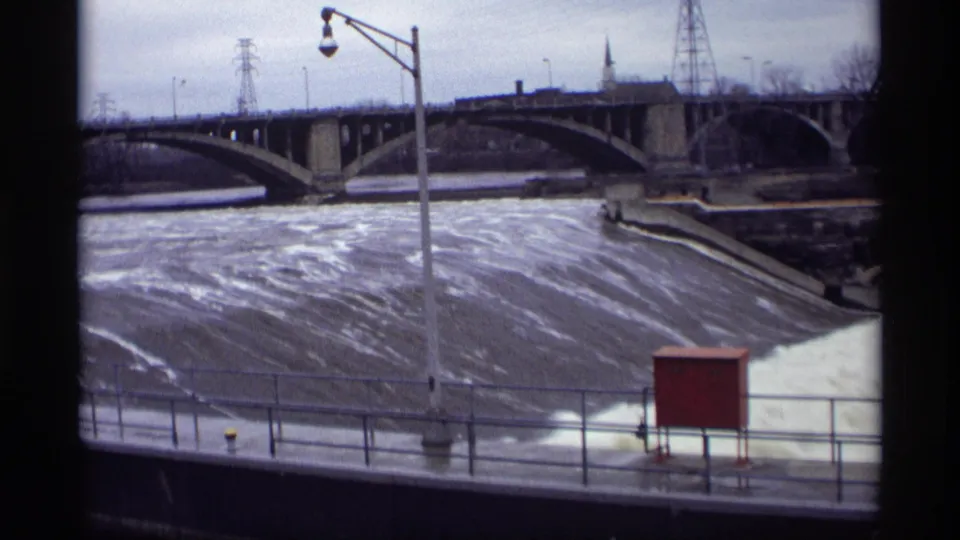 A 1969 8mm view shows turbulent water at St. Anthony Falls with a concrete bridge, railing and power structures in the background.