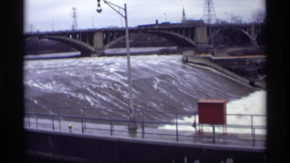 The fixed 8mm frame looks across St. Anthony Falls toward a concrete bridge, churning water, utility lines and a red riverside box.