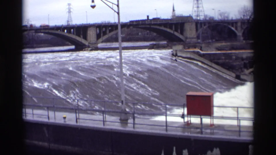 A final 1969 8mm frame holds on the St. Anthony Falls spillway with turbulent water, a bridge, railings and power towers.