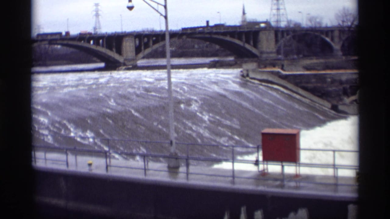 Concrete spillway with rushing water, railing, red utility box, and arched bridge beyond.