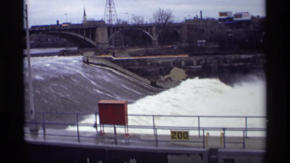 1969 Savage, Minnesota view of turbulent water at the dam edge, with a foreground railing, yellow 200 sign, red utility box, and arched bridge.