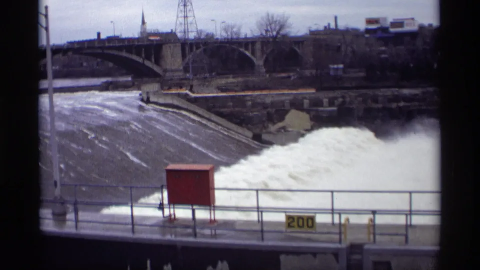 1969 Savage, Minnesota 8mm frame with foaming water at a dam, a red utility box in front, metal railing, bridge spans, and riverbank structures.