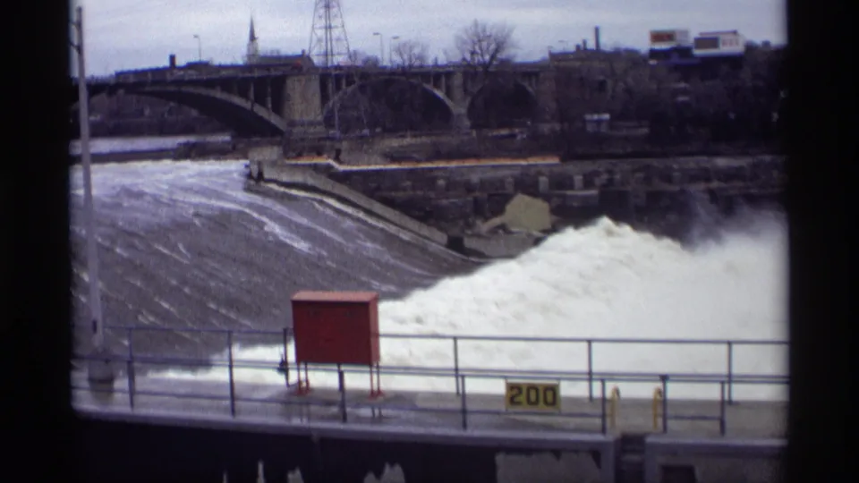 1969 Savage, Minnesota home-movie frame showing a fixed view of the dam spillway, heavy white water, foreground railing, red utility box, and bridge.