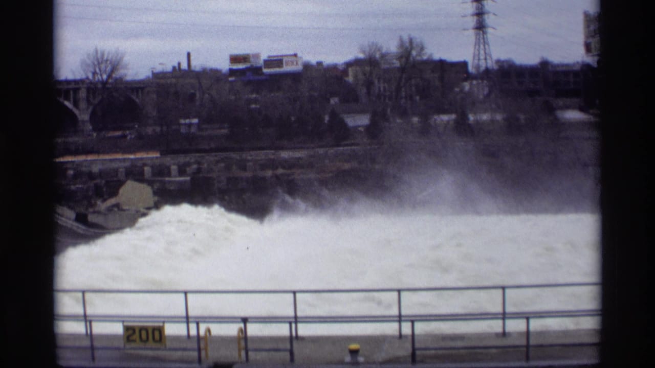 Foaming water rushes over a dam behind a metal railing and yellow 200 sign.