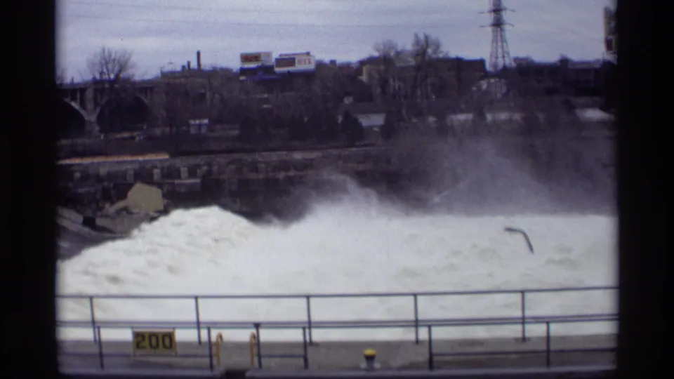 Savage, Minnesota dam footage shows turbulent white water and spray, with a walkway railing, distant trees, buildings, and power structures visible.