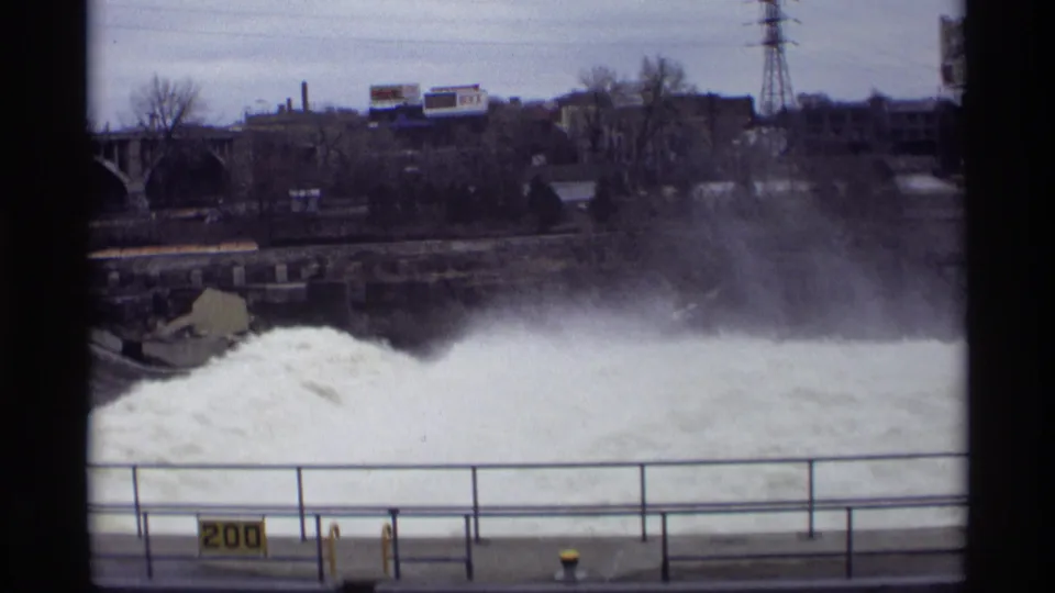 A 1969 home-movie frame holds on rushing dam water in Savage, with the railing, yellow sign, treeline, bridge edge, and industrial background visible.