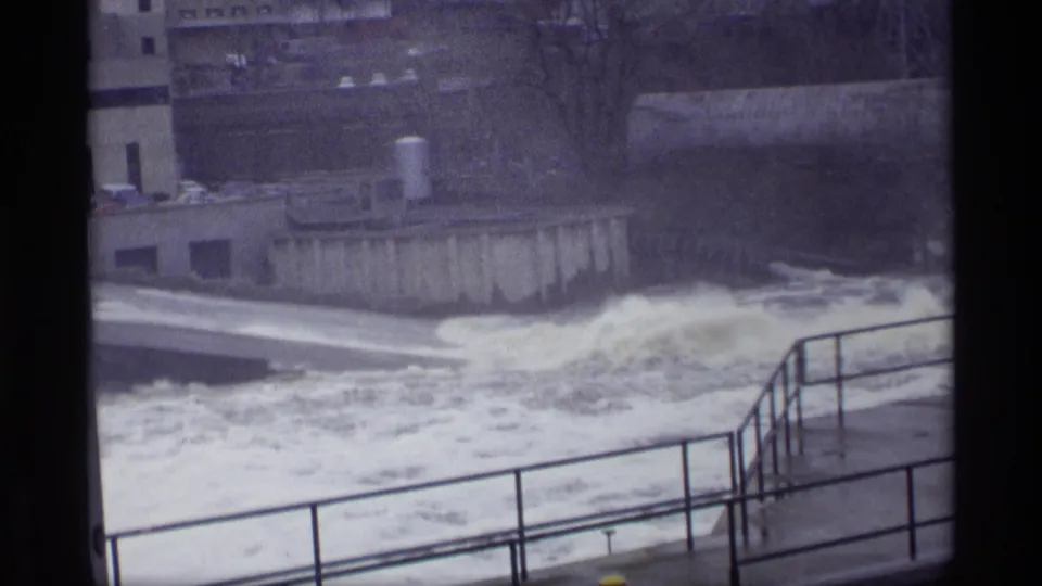 1969 8mm view of turbulent water at a concrete dam spillway in Savage, Minnesota, seen from behind metal railings.