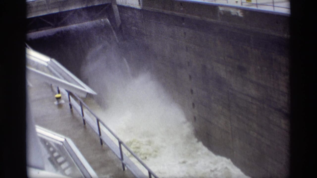 Turbulent water and spray rushing through a concrete dam spillway beside railings and a walkway.