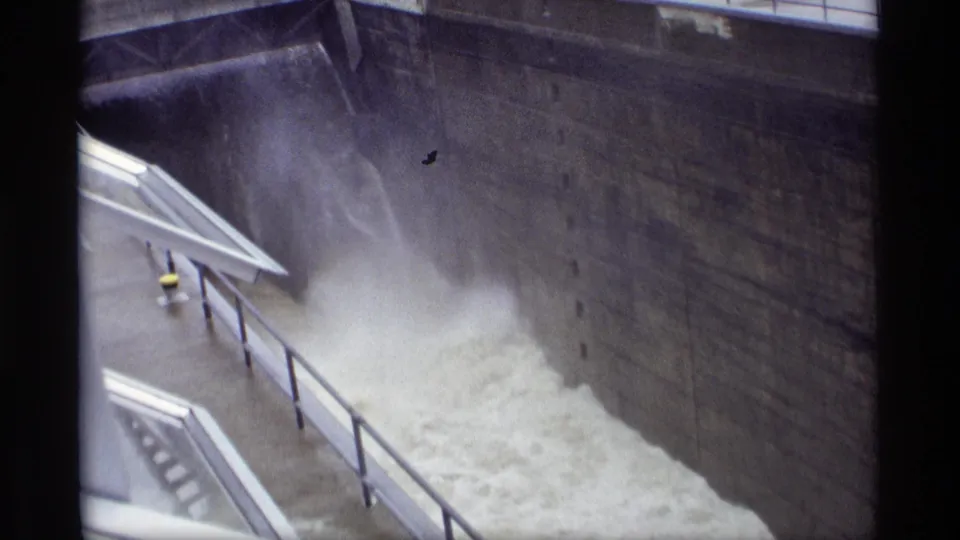 1969 Savage, Minnesota dam footage showing foaming spillway water below a tall concrete wall, with railings, walkway edges, and film specks visible.