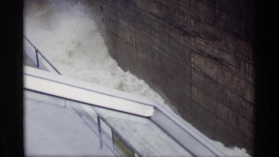 Home-movie frame from the 1969 Savage, Minnesota dam scene, with rushing water, concrete wall, stairs, railings, and walkway edges in view.