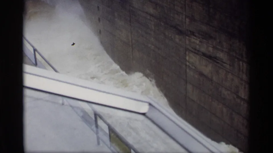 1969 8mm Savage, Minnesota dam frame showing mist, foaming water, a concrete wall, foreground walkway, railings, and dark film borders.
