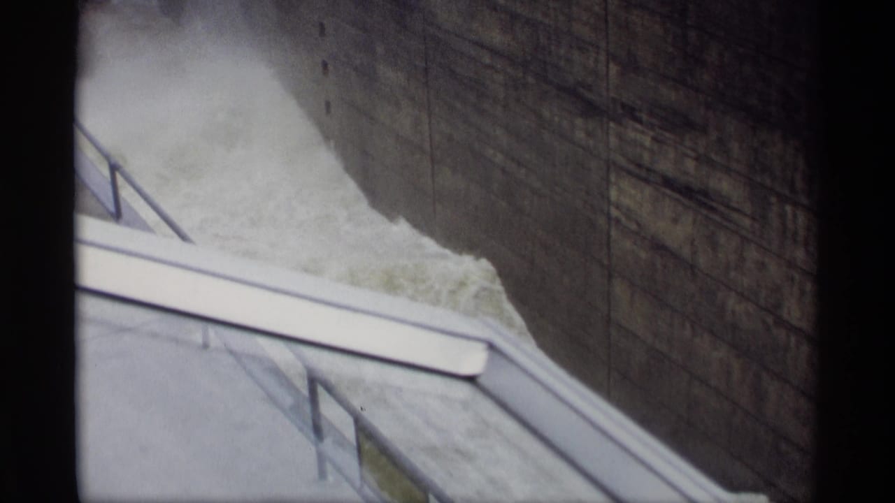 Turbulent water beside a concrete wall and sloped railing under a bridge shadow.